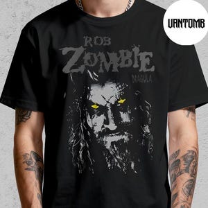 Puede incluir: Camiseta negra con un retrato en escala de grises de Rob Zombie y el texto "ROB ZOMBIE" encima de la imagen y "MAGNOLIA" debajo. El retrato tiene ojos amarillos. La camiseta la lleva una persona.