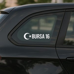 Op de afbeelding: Witte sticker met een maan en ster symbool, met de tekst "BURSA 16" in een schreefloos lettertype. De sticker is bevestigd op een zwarte autoruit.