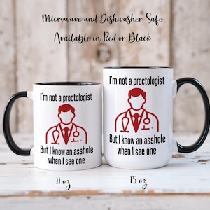 Op de afbeelding: Twee witte keramische mokken met zwarte handvatten en randen. Elke mok heeft de tekst "I'm not a proctologist But I know an asshole when I see one" en een rode afbeelding. De mokken zijn gelabeld met 11 oz en 15 oz.
