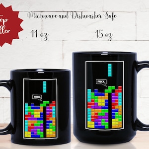Puede incluir: Dos tazas de cerámica negra en tamaños de 11 oz y 15 oz. Cada taza presenta un gráfico rectangular de un videojuego de bloques de colores con un globo de texto que contiene la palabra FUCK. Las tazas son aptas para microondas y lavavajillas.