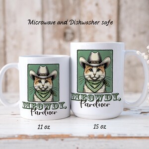 May include: Two white ceramic mugs featuring a cartoon cat wearing a cowboy hat and bandana. The mugs read "MEOWDY, Pardner". The smaller mug is 11 oz and the larger mug is 15 oz.