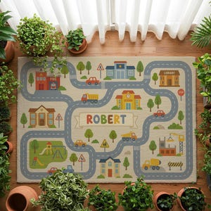 Puede incluir: Alfombra de juego beige para niños con diseño de mapa de carreteras. La alfombra presenta carreteras, edificios como una estación de bomberos, una escuela y una biblioteca, y el nombre "ROBERT" en una pancarta. También se representan coches, camiones y árboles.