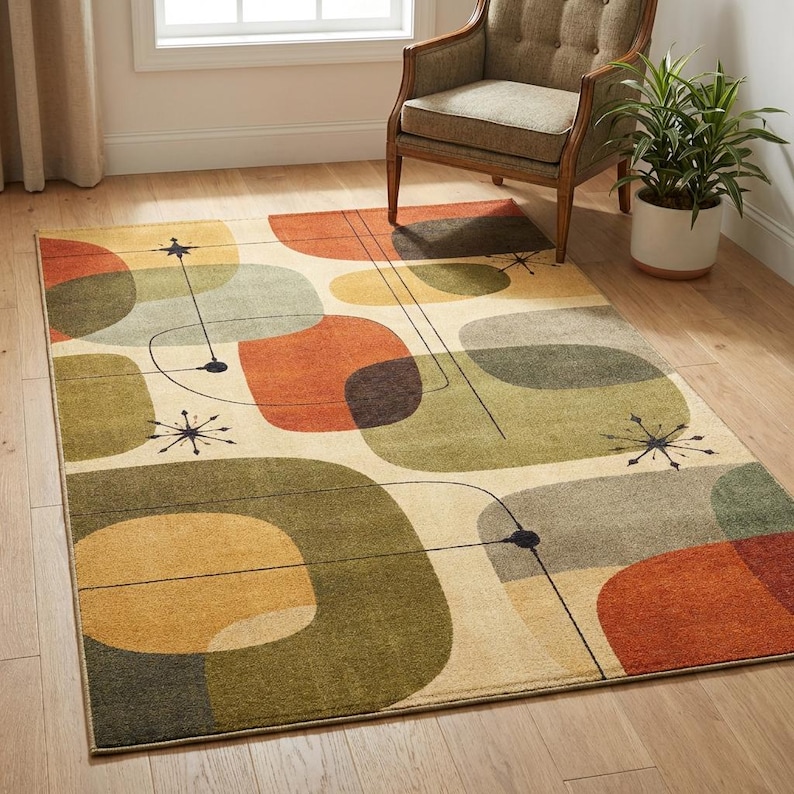 May include: A rectangular area rug with an abstract mid-century modern design. The rug features large, overlapping shapes in shades of orange, green, and beige, with black lines and starburst accents. The rug is placed on a hardwood floor.