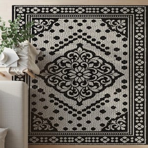 May include: A black and white patterned area rug with a floral medallion design. The rug features a border with geometric and floral patterns. The central design is a large, ornate floral motif. The rug is placed on a light-colored wooden floor.
