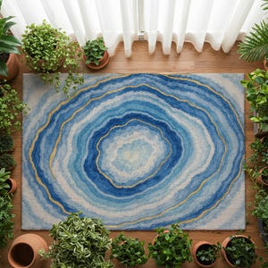 Könnte beinhalten: Ein rechteckiger Teppich mit einem blau-weißen Achat-Design und goldenen Akzenten. Der Teppich ist von Topfpflanzen auf einem Holzboden umgeben, mit weißen Vorhängen im Hintergrund. Das Design des Teppichs ähnelt konzentrischen Kreisen.