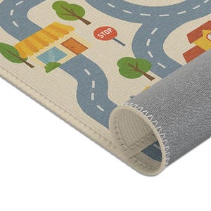 Puede incluir: Alfombra de juego beige con un dise&ntilde;o de carretera que presenta carreteras azules, &aacute;rboles verdes y edificios con tejados amarillos. Es visible una se&ntilde;al de stop roja. La alfombra est&aacute; enrollada en la parte inferior, revelando un respaldo gris.