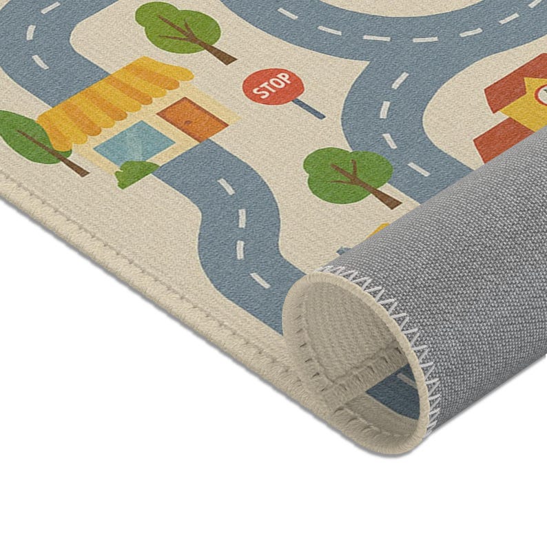 Puede incluir: Alfombra de juego beige con un dise&ntilde;o de carretera que presenta carreteras azules, &aacute;rboles verdes y edificios con tejados amarillos. Es visible una se&ntilde;al de stop roja. La alfombra est&aacute; enrollada en la parte inferior, revelando un respaldo gris.