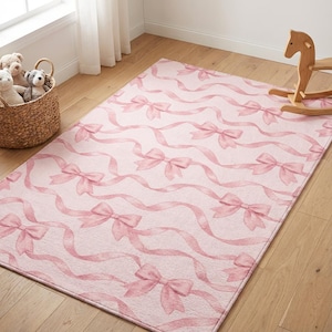Può includere: Tappeto rettangolare rosa con un motivo ripetuto di fiocchi e nastri rosa. Il tappeto è posizionato su un pavimento in legno, con un cestino di vimini con peluche e un cavallo a dondolo in legno nelle vicinanze.