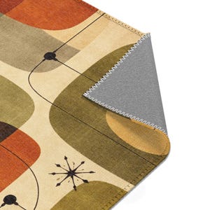 May include: A mid-century modern area rug with an abstract design. The rug features a cream background with orange, olive green, and brown geometric shapes. Black lines and starburst details add to the retro aesthetic. The underside of the rug is gray.