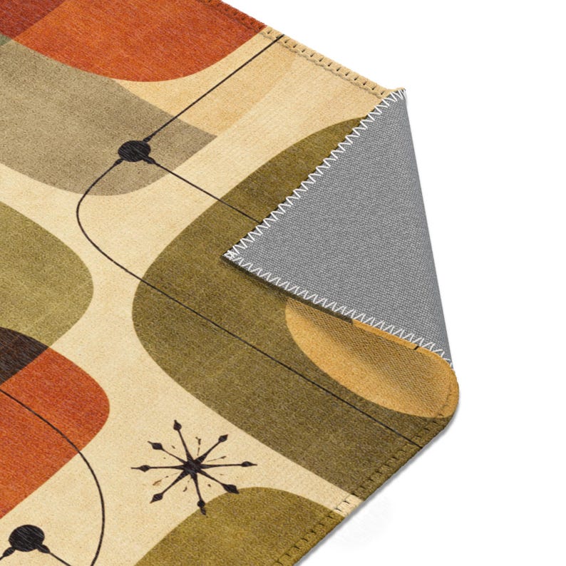 May include: A mid-century modern area rug with an abstract design. The rug features a cream background with orange, olive green, and brown geometric shapes. Black lines and starburst details add to the retro aesthetic. The underside of the rug is gray.