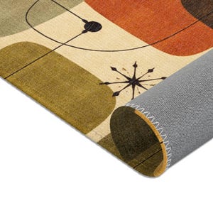 May include: A rolled-up area rug with a mid-century modern design. The rug features a beige background with olive green, orange, and brown geometric shapes. Black lines and starburst details add to the retro aesthetic.