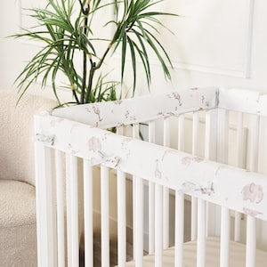 Puede incluir: Cuna blanca con un protector suave estampado con elefantes y jirafas en tonos marrón y beige. El protector está atado a la cuna con pequeños lazos de color claro. Una planta verde está en el fondo.