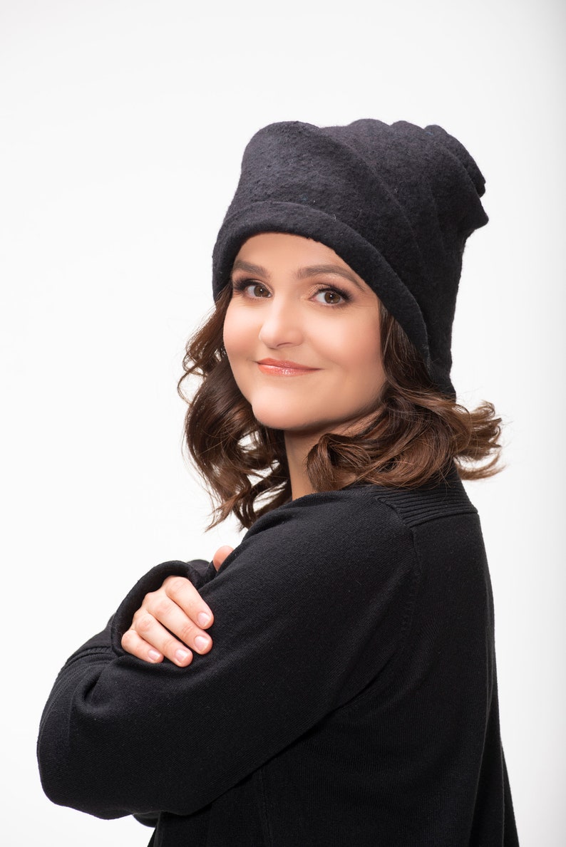 May include: A woman with brown hair wearing a black beanie and a black sweater. She is looking at the camera and has her arms crossed.