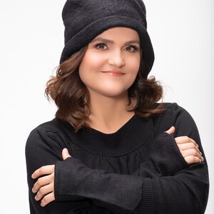 May include: A woman with brown hair wearing a black beanie and a black sweater with her arms crossed.