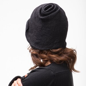 May include: A black felt hat with a unique rose-like design. The hat is worn by a person with brown hair, but the face is not visible.