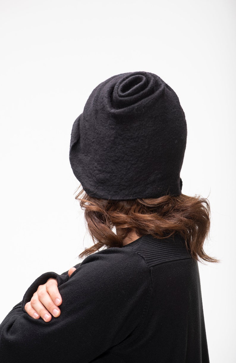 May include: A black felt hat with a unique rose-like design. The hat is worn by a person with brown hair, but the face is not visible.