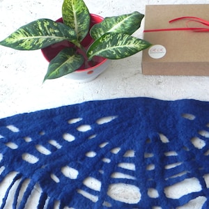 May include: A vibrant blue felt scarf with a unique cut-out design and fringe detail. The scarf is displayed with a potted plant and a brown gift box tied with a red ribbon. The background is a textured white surface.