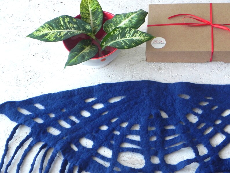 May include: A vibrant blue felt scarf with a unique cut-out design and fringe detail. The scarf is displayed with a potted plant and a brown gift box tied with a red ribbon. The background is a textured white surface.