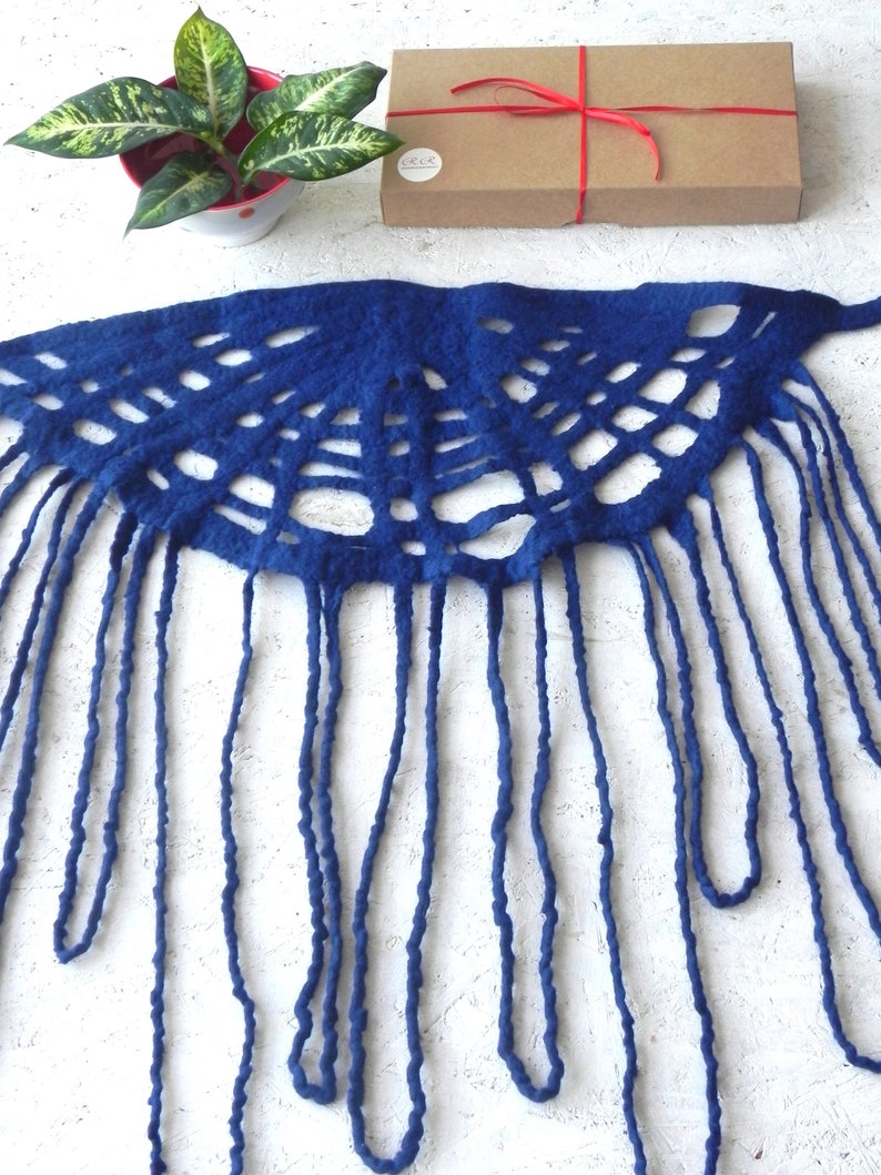 May include: A deep blue, handmade felt scarf with a unique openwork design and long fringe. The scarf is displayed on a white surface, with a small potted plant and a brown gift box tied with red ribbon in the background.