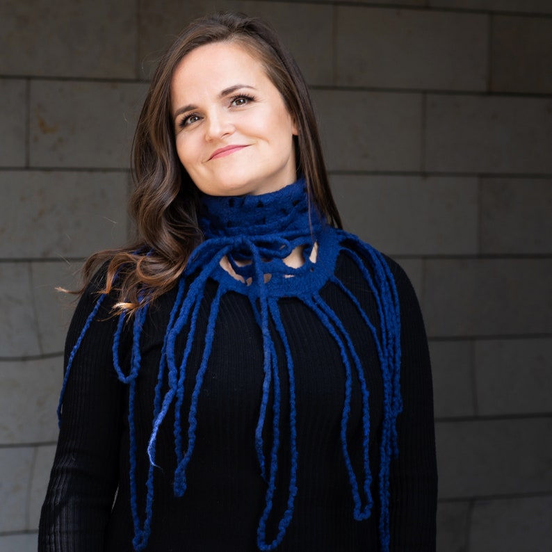 May include: A woman wearing a dark blue, handmade felted cowl neck warmer. The cowl has a high collar with a decorative openwork design and long, dangling fringe. The woman is wearing a black long-sleeved shirt.