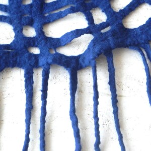 May include: Close-up of a handmade, royal blue felt textile. The felt has a cut-out pattern with irregular shapes and long, dangling fringe. The texture of the felt is visible against a white background.
