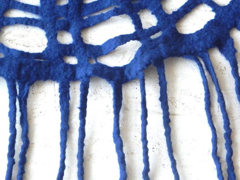 May include: Close-up of a handmade, royal blue felt textile. The felt has a cut-out pattern with irregular shapes and long, dangling fringe. The texture of the felt is visible against a white background.