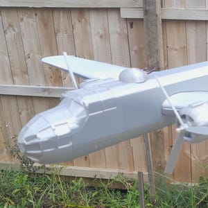 May include: A silver model airplane with a propeller, wings, and a tail. The model is sitting on a wooden fence.
