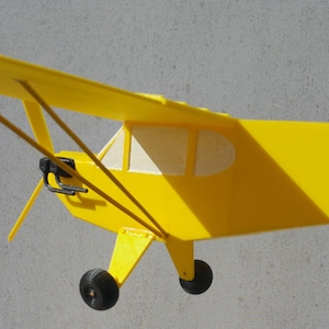 Puede incluir: Un avión modelo amarillo con hélice y tren de aterrizaje. El avión tiene una ventana blanca en el lateral.