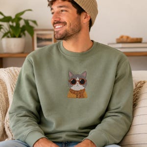 May include: An olive green crewneck sweatshirt featuring a central embroidered graphic of a gray and white cat wearing round orange-rimmed sunglasses and a mustard yellow plaid scarf over a matching buttoned cardigan.