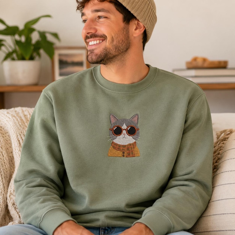 May include: An olive green crewneck sweatshirt featuring a central embroidered graphic of a gray and white cat wearing round orange-rimmed sunglasses and a mustard yellow plaid scarf over a matching buttoned cardigan.