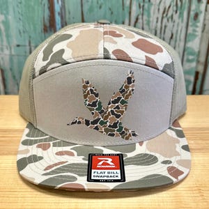 May include: A flat bill snapback hat with a camouflage pattern in shades of brown, tan, and green. The front panel features a duck graphic in a camouflage design. The hat has a mesh back and a "Richardson" patch.