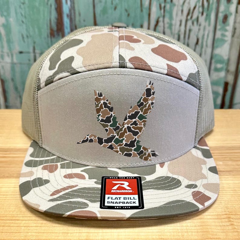 May include: A flat bill snapback hat with a camouflage pattern in shades of brown, tan, and green. The front panel features a duck graphic in a camouflage design. The hat has a mesh back and a "Richardson" patch.