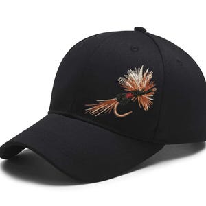 May include: Black baseball cap with a curved brim. The cap features an embroidered fly fishing lure design in shades of brown, white, and red. The cap has a button on top and is made of a durable fabric.