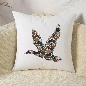 May include: White square throw pillow featuring a flying duck silhouette. The duck's wings and body are filled with a camouflage pattern in shades of brown, green, and black. The pillow is on a neutral-colored surface.
