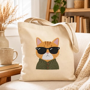 May include: A beige canvas tote bag featuring an embroidered graphic of an orange tabby cat wearing black sunglasses and a dark green button-up sweater. The cat has a white muzzle, pink inner ears, and a serious expression on its face.