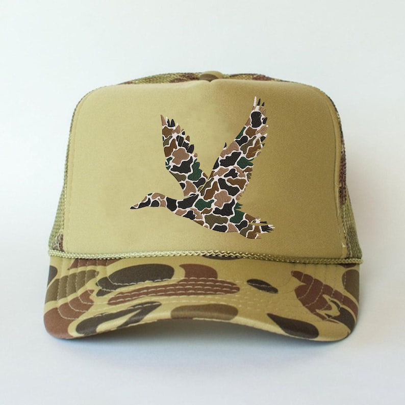 May include: A tan trucker hat with a camouflage duck graphic. The hat features a mesh back, a curved brim with a camouflage pattern, and a tan front panel with a flying duck design in shades of brown, green, and black.