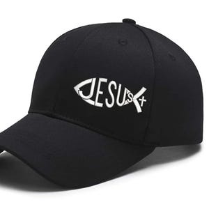 May include: Black baseball cap featuring the word "JESUS" in white, designed within a fish symbol. The cap has a curved brim and a button on top. The hat is made of a durable fabric.