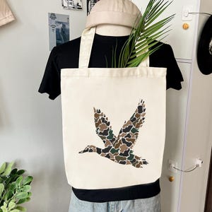 May include: Cream-colored canvas tote bag featuring a flying duck design in camouflage pattern. The bag has a rectangular shape with two handles. The duck design incorporates shades of brown, green, and black.