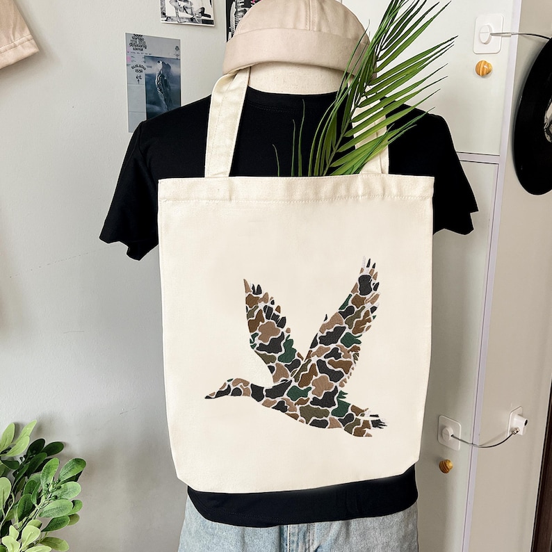 May include: Cream-colored canvas tote bag featuring a flying duck design in camouflage pattern. The bag has a rectangular shape with two handles. The duck design incorporates shades of brown, green, and black.