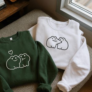 Könnte beinhalten: Zwei Sweatshirts, eines in Dunkelgrün und eines in Weiß, werden gezeigt. Jedes Sweatshirt hat ein Cartoon-Frosch-Design. Das grüne Sweatshirt hat eine Herzgrafik. Das weiße Sweatshirt hat ein ähnliches Frosch-Design. Beide sind aus weichem Material.