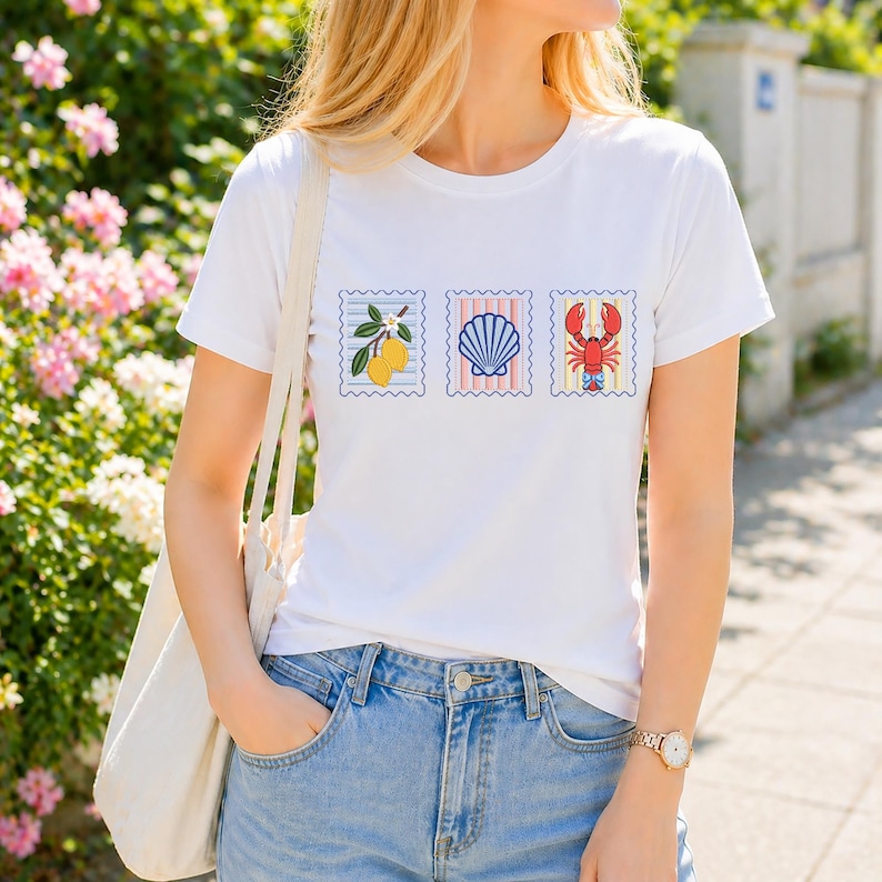 May include: A white short-sleeved t-shirt featuring three rectangular postage stamp graphics arranged horizontally. The designs include a yellow lemon branch, a blue seashell, and a red lobster, each framed by a scalloped border.
