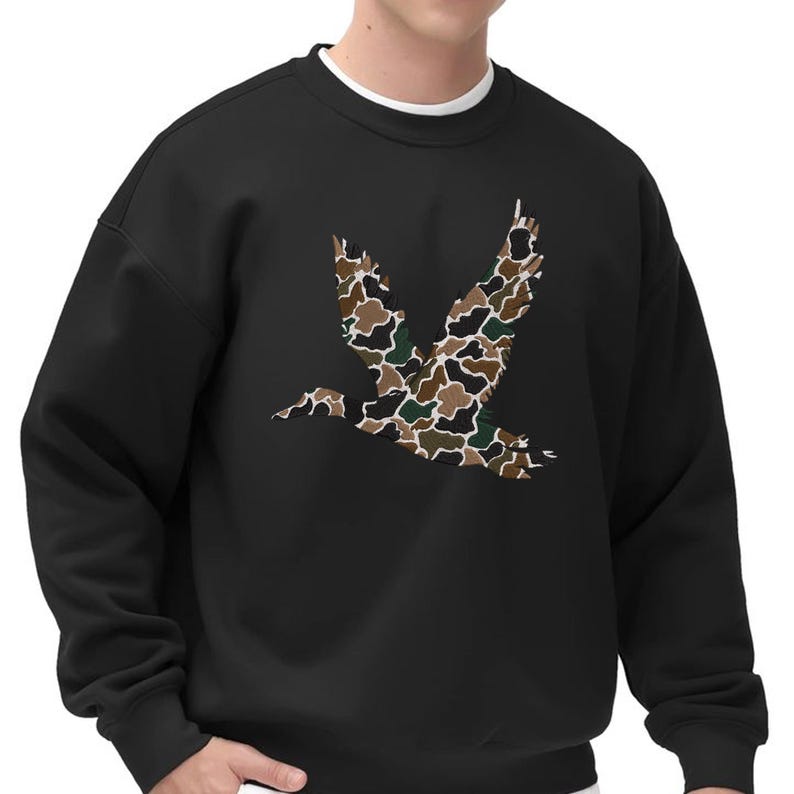 May include: Black sweatshirt featuring a large, stylized bird in flight. The bird is filled with a camouflage pattern in shades of brown, green, and white. The sweatshirt has a crew neck with a white undershirt visible at the neckline.