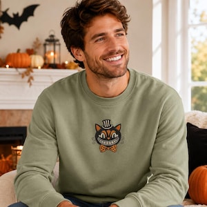 Puede incluir: Una sudadera de cuello redondo en color verde salvia con un gr&aacute;fico bordado central de un gato negro con sombrero de copa y pajarita naranja de lunares. El gato tiene una amplia sonrisa, ojos naranjas y bigotes estilizados en el pecho.