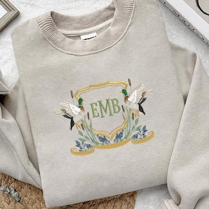 May include: Beige sweatshirt with a crew neck and embroidered crest. The crest features two ducks, reeds, and the letters "EMB" in green. A gold-colored banner and frame surround the design. The sweatshirt is a neutral color.