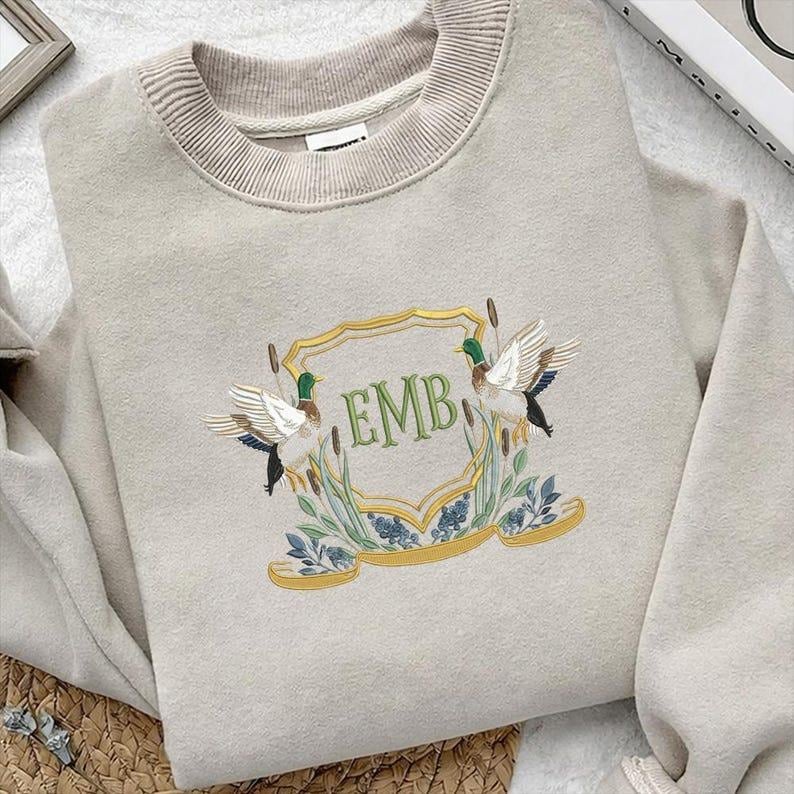 May include: Beige sweatshirt with a crew neck and embroidered crest. The crest features two ducks, reeds, and the letters "EMB" in green. A gold-colored banner and frame surround the design. The sweatshirt is a neutral color.