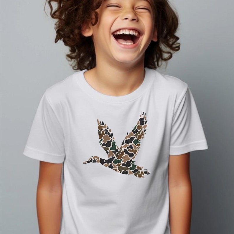 May include: White t-shirt featuring a camouflage duck graphic in shades of brown, green, and black. The short-sleeved shirt is worn by a child. The duck is in mid-flight, with its wings spread wide.