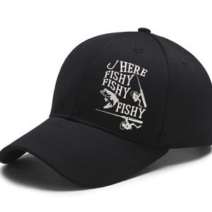 May include: Black baseball cap with the text "HERE FISHY FISHY FISHY" in white. The design includes a fish, fishing rod, and reel. The cap has a curved brim and a button on top.