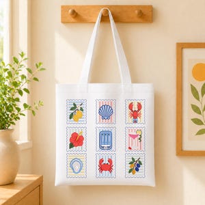 May include: A white tote bag featuring a grid of nine postage stamp-style illustrations. The designs include lemons, a seashell, a lobster, a hibiscus flower, a tin of sardines, a cocktail, an oyster, a crab, and olives on a branch.