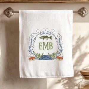 May include: White hand towel with a fishing-themed design. The towel features a central emblem with a fish, fishing rod, and the initials "EMB" in green. The design includes blue and green accents, with two small tackle boxes.