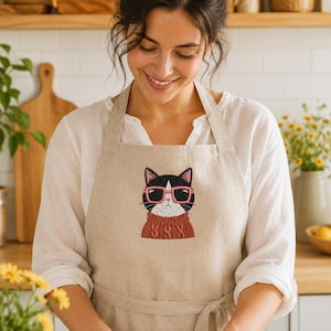 May include: A beige linen apron featuring an embroidered graphic of a black and white cat wearing pink-rimmed glasses and a textured rust-colored cable knit sweater. The apron has a simple neck loop and waist ties for a secure fit.